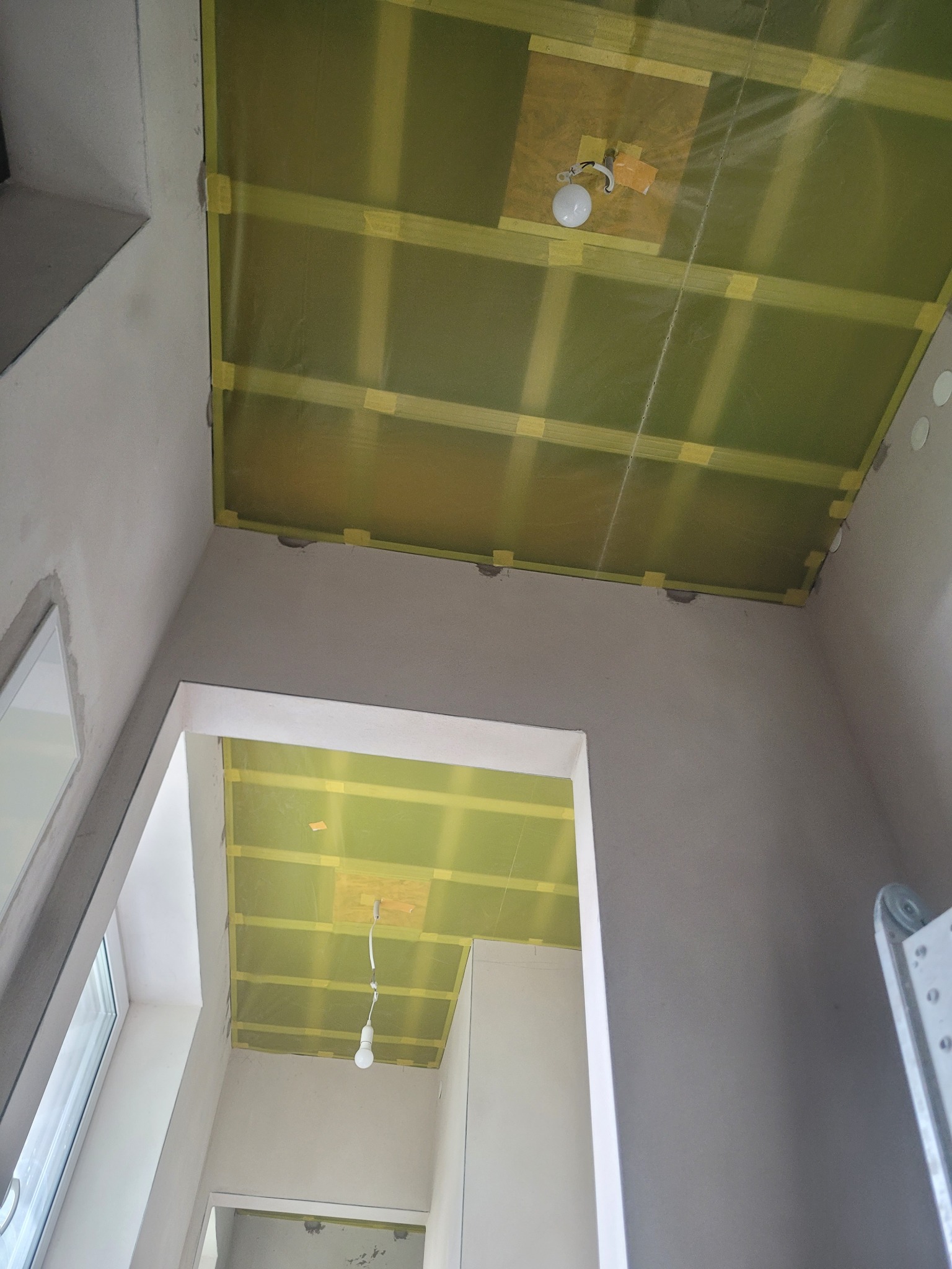 Sufit podwieszany w trakcie montażu: widoczne profile, folia paroizolacyjna i taśma klejąca. W tle fragment okna i drabiny. Ujęcie z dołu.