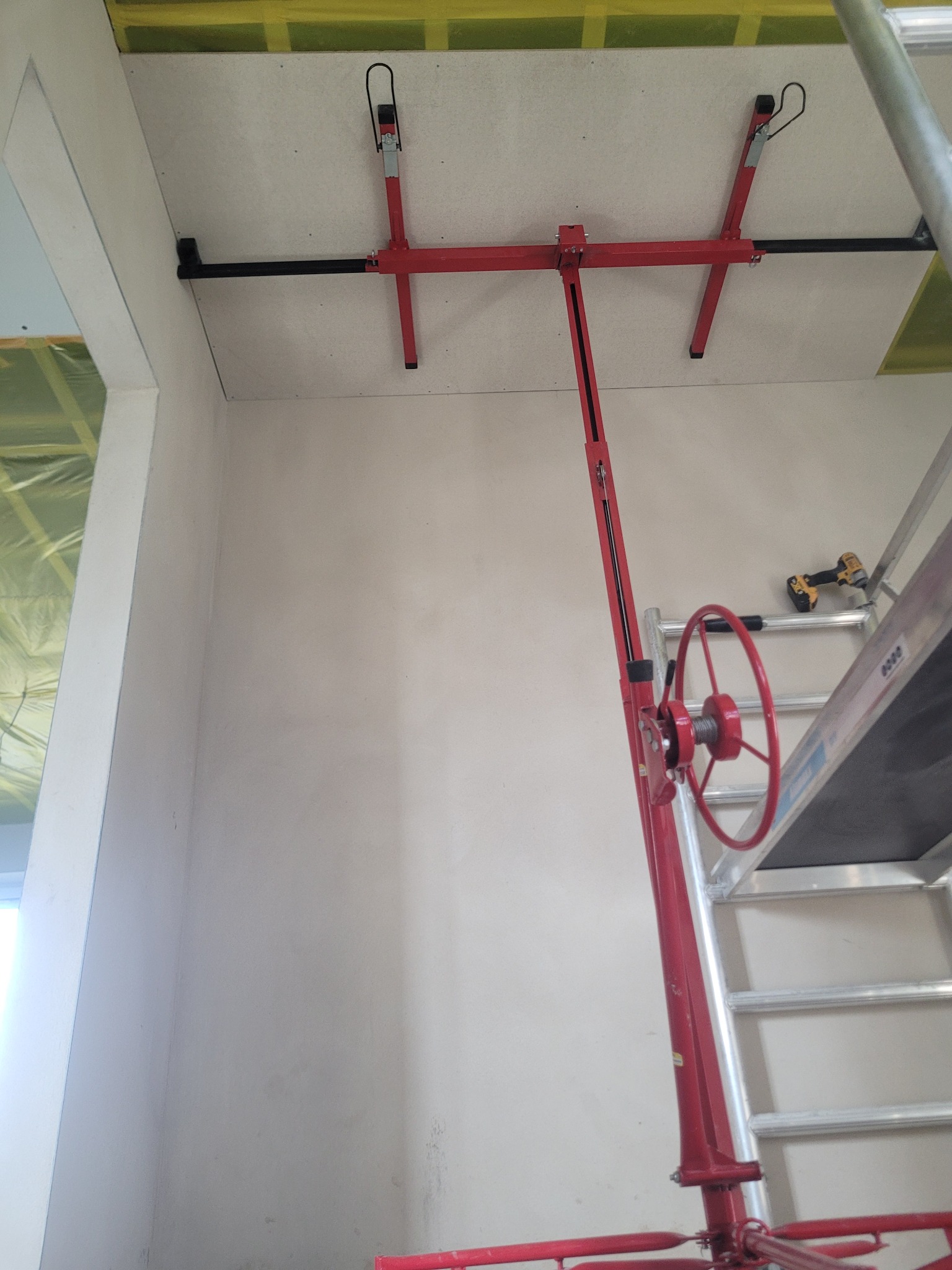 Montaż płyty gipsowej na suficie za pomocą podnośnika. Widoczny fragment rusztowania i ściana z gładzią. Żółta folia w tle.