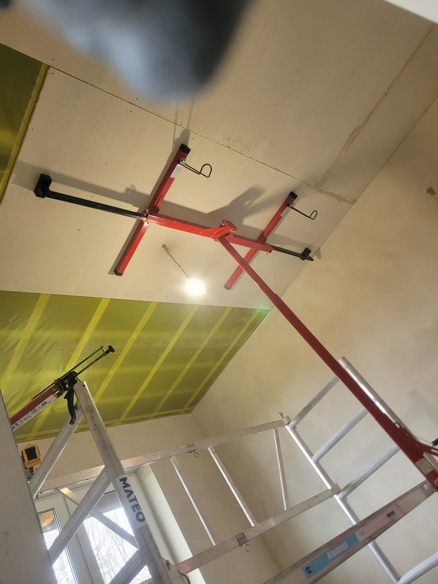 Montaż płyty gipsowej na suficie w Kamionnej z użyciem podnośnika. Widoczna drabina i narzędzia. Żółta izolacja za płytą. Ujęcie z dołu.