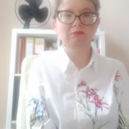 Portret kobiety w okularach i białej koszuli w kwiaty, siedzącej na krześle biurowym na tle białego regału z książkami i czarnym wentylatorem na górze.