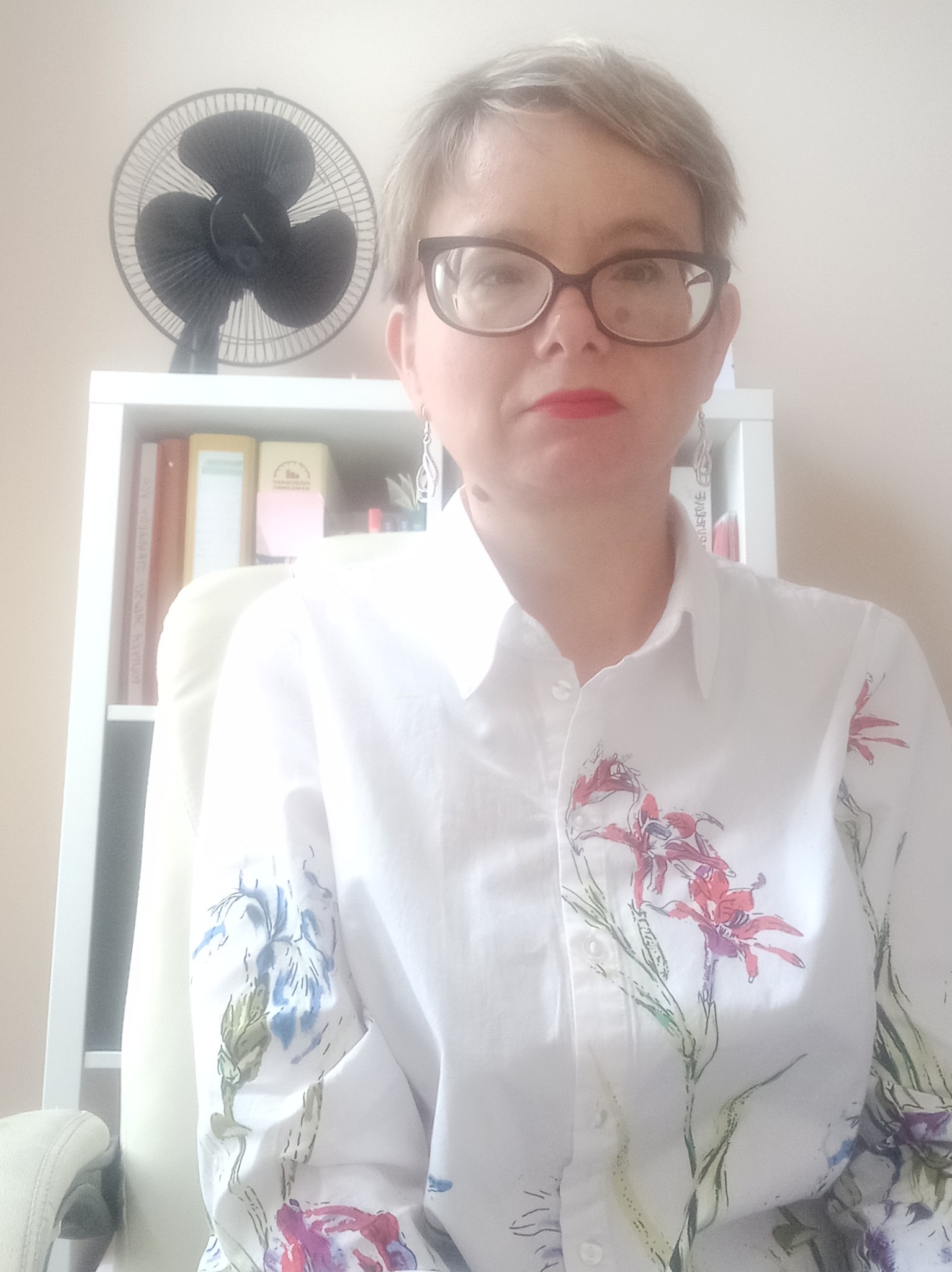 Portret kobiety w okularach i białej koszuli w kwiaty, siedzącej na krześle biurowym na tle białego regału z książkami i czarnym wentylatorem na górze.