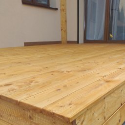 VIKING ŁUKASZ ZIMKOWSKI - Nowoczesny, jasny taras z desek drewnianych, przylegający do budynku z jasną elewacją i ciemnymi oknami, widok z niskiej perspektywy ukazujący strukturę drewna.