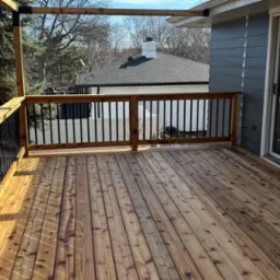 Nowy taras z desek z drewna, czarne metalowe balustrady, widok na dom z szarą elewacją i białą balustradą w tle.