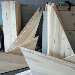 VIKING ŁUKASZ ZIMKOWSKI - Elementy schodów drewnianych z jasnego drewna o widocznych słojach, przygotowane do montażu, składowane w pomieszczeniu z szaro-białymi płytkami na ścianie.