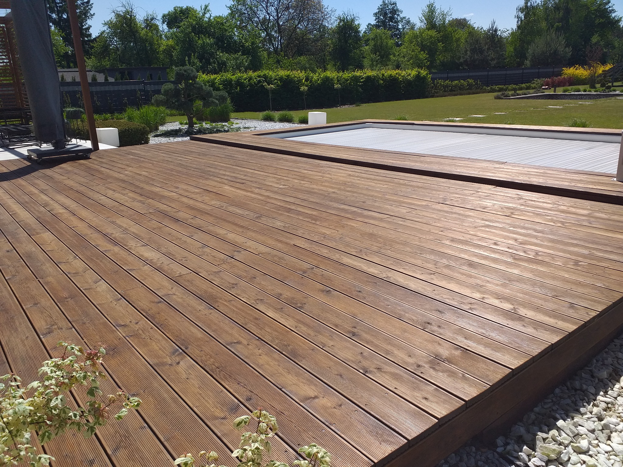 Nowoczesny taras z desek kompozytowych w odcieniach brązu, otoczony białymi kamieniami i zielenią ogrodu, z widocznym zadaszeniem basenu w tle.