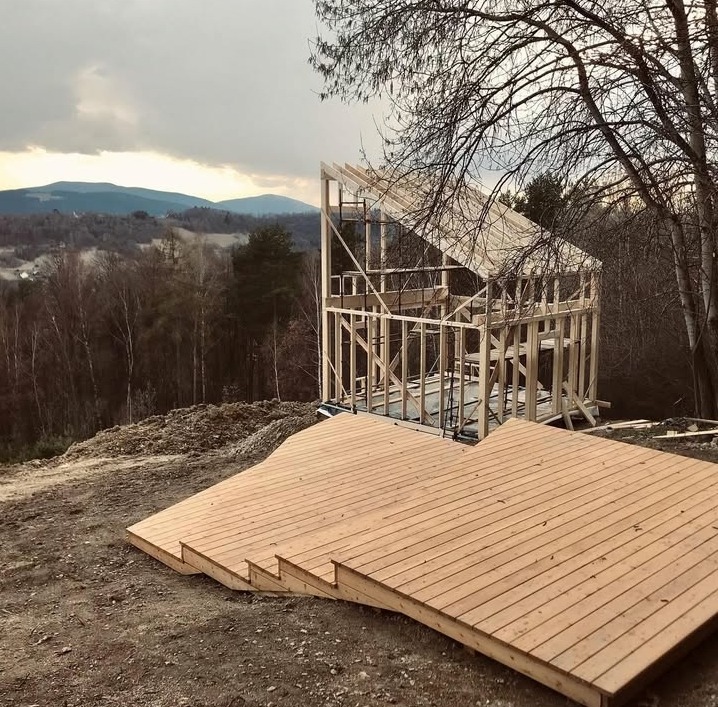 Wznoszenie domu w konstrukcji szkieletowej na wzgórzu, z widocznym drewnianym tarasem w trakcie budowy i panoramą górską w tle, pochmurne niebo.