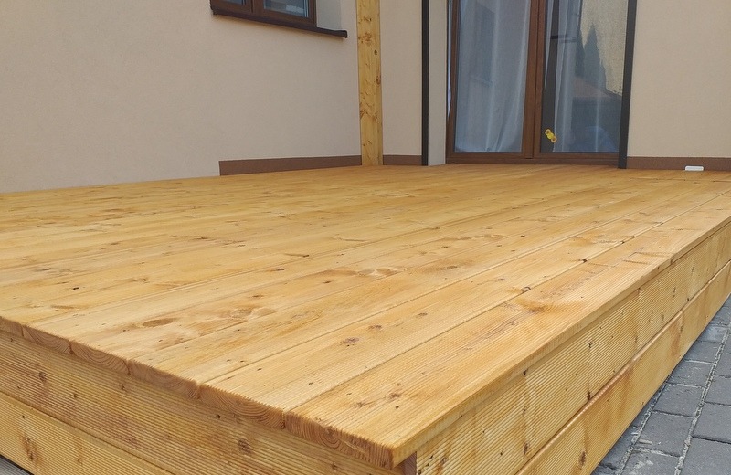 Nowoczesny, jasny taras z desek drewnianych, przylegający do budynku z jasną elewacją i ciemnymi oknami, widok z niskiej perspektywy ukazujący strukturę drewna.