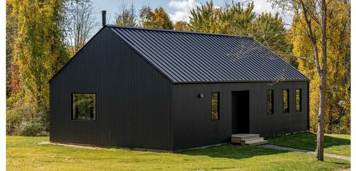 Czarny, nowoczesny dom z metalowym dachem, otoczony zielenią i drzewami w jesiennej scenerii.