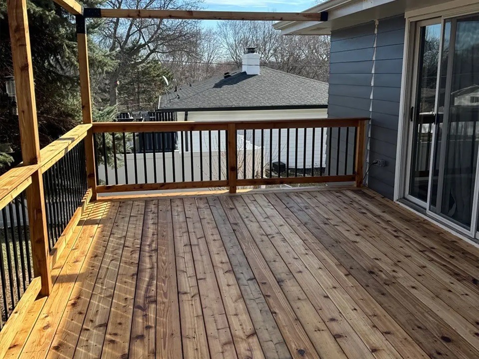 Nowy taras z desek z drewna, czarne metalowe balustrady, widok na dom z szarą elewacją i białą balustradą w tle.