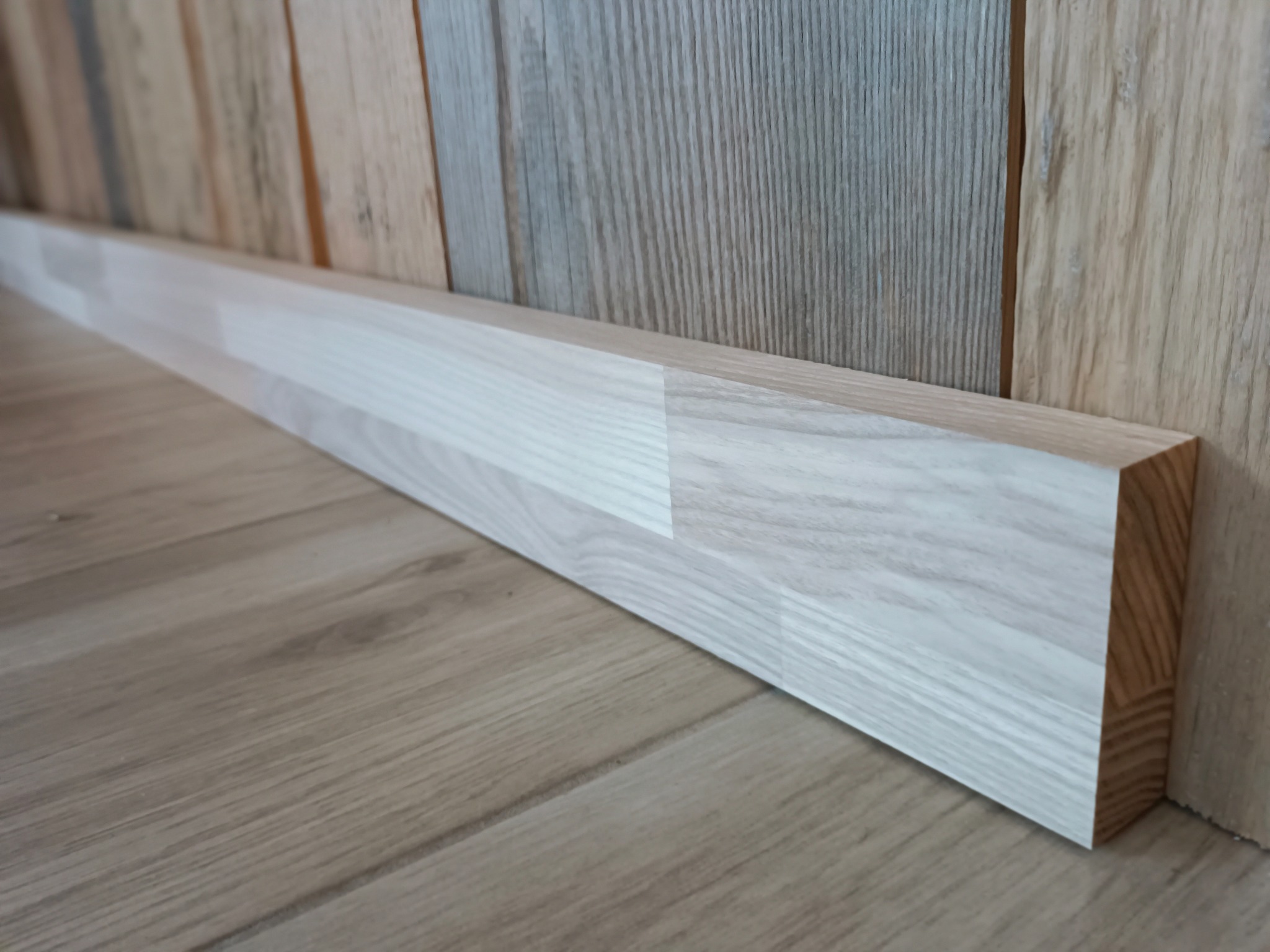 Zamontowana, jasna listwa przypodłogowa z drewna na styku drewnianej podłogi i ściany wyłożonej panelami o zróżnicowanej kolorystyce drewna.