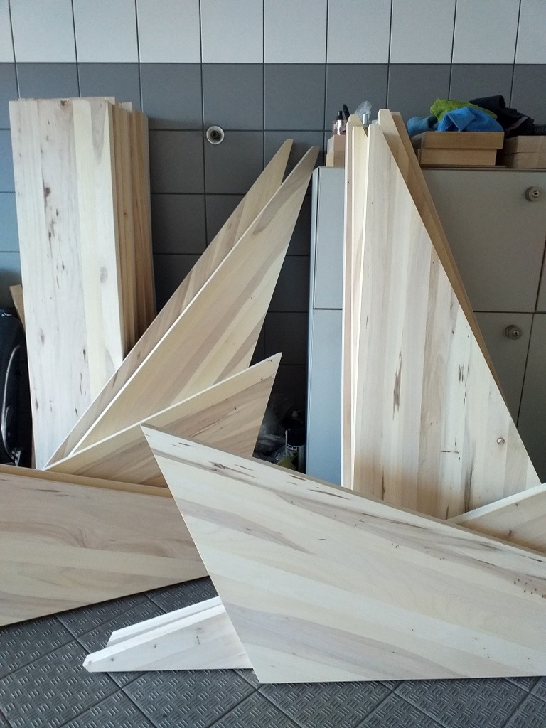 Elementy schodów drewnianych z jasnego drewna o widocznych słojach, przygotowane do montażu, składowane w pomieszczeniu z szaro-białymi płytkami na ścianie.