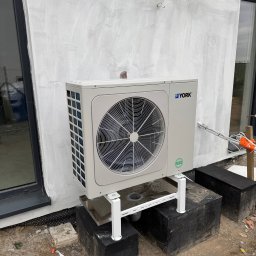 Pro-Inst - Zewnętrzna jednostka klimatyzacji marki York zamontowana na białym stelażu, przy ścianie budynku z surowym tynkiem, widoczne okno i przewody w izolacji.