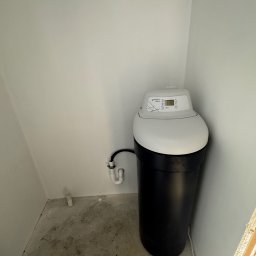 Pro-Inst - Czarno-biały zmiękczacz wody marki Trinity Home 2.0 zainstalowany w rogu pomieszczenia z surowym betonem na podłodze i widocznymi rurami przyłączeniowymi do ściany.