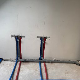 Pro-Inst - Wewnętrzna instalacja wodna z rurami PEX w kolorze niebieskim i czerwonym, wychodzącymi ze ściany, widoczne mocowania i szare rury kanalizacyjne, surowy stan wykończenia wnętrza.