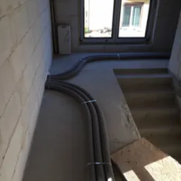 Szare, karbowane rury wentylacyjne prowadzone wzdłuż ściany z betonu komórkowego w surowym stanie, zabezpieczone metalowymi opaskami, z widocznym oknem i schodami w tle.