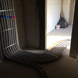 Pro-Inst - Szare rury instalacyjne, zabezpieczone metalowymi taśmami, prowadzone w surowym wnętrzu budynku z bloczków, widoczne oznaczenia kolorami niebieskim i czerwonym.