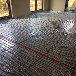 Pro-Inst - Białe rurki ogrzewania podłogowego ułożone w pętle na czarnej siatce, zabezpieczone folią, z widocznymi czerwonymi taśmami mocującymi w jasnym pomieszczeniu z dużymi oknami.