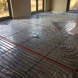 Białe rurki ogrzewania podłogowego ułożone w pętle na czarnej siatce, zabezpieczone folią, z widocznymi czerwonymi taśmami mocującymi w jasnym pomieszczeniu z dużymi oknami.