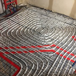 Pro-Inst - Białe rury systemu ogrzewania podłogowego ułożone spiralnie na czarnej siatce, przymocowane plastikowymi klipsami, z czerwonymi taśmami wyznaczającymi strefy, widoczne fragmenty instalacji...