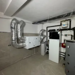 Zainstalowana jednostka rekuperacyjna z aluminiowymi kanałami wentylacyjnymi w pomieszczeniu technicznym z szarą płytką podłogową i kotłem grzewczym po prawej stronie.