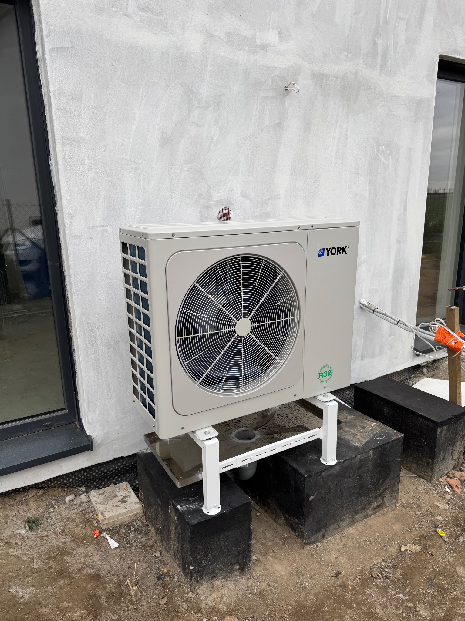 Zewnętrzna jednostka klimatyzacji marki York zamontowana na białym stelażu, przy ścianie budynku z surowym tynkiem, widoczne okno i przewody w izolacji.