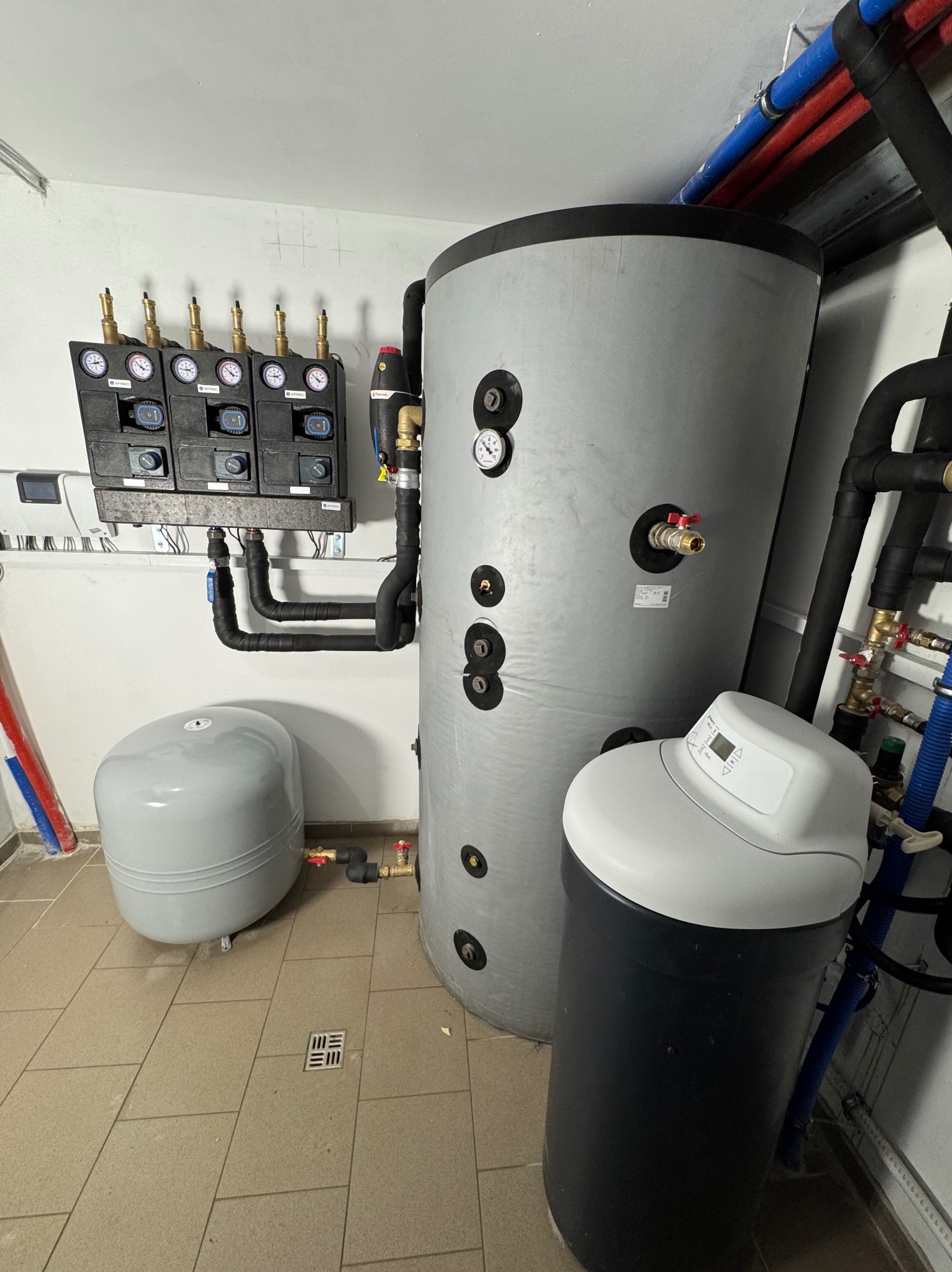 Szara instalacja z zbiornikiem wody, naczyniem przeponowym i stacją uzdatniania wody w kotłowni, z czarnymi rurami i manometrami na białej ścianie.