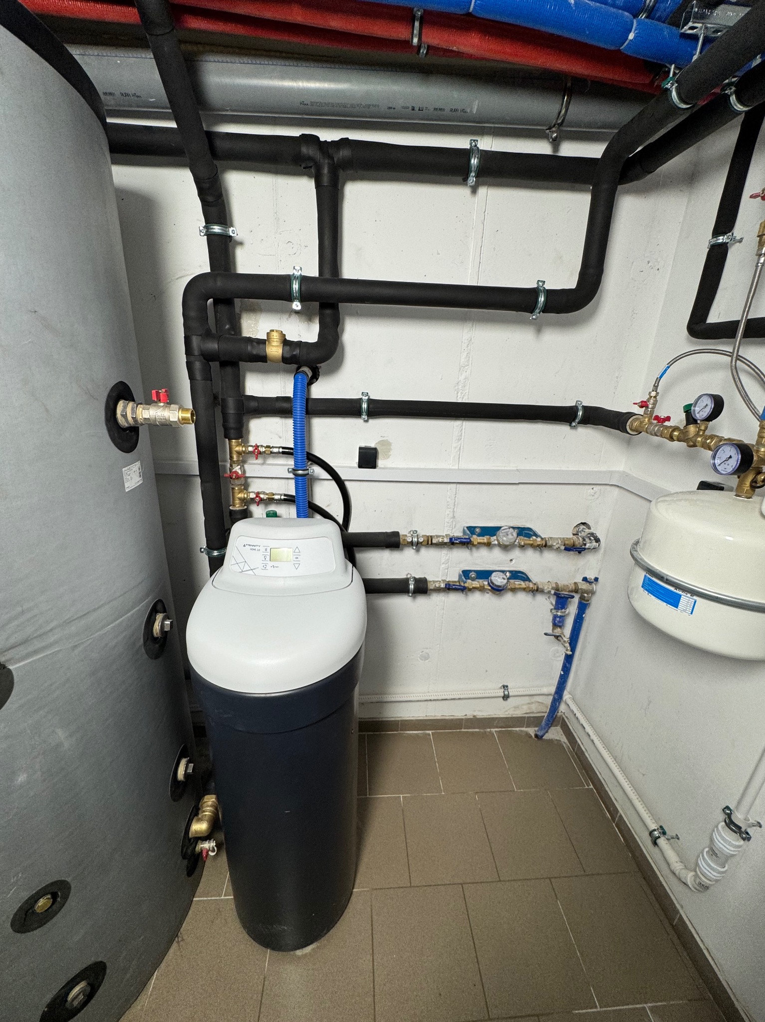 Szara instalacja z rurami w izolacji, zbiornik buforowy i zmiękczacz wody z wyświetlaczem, widoczne liczne zawory i manometry.