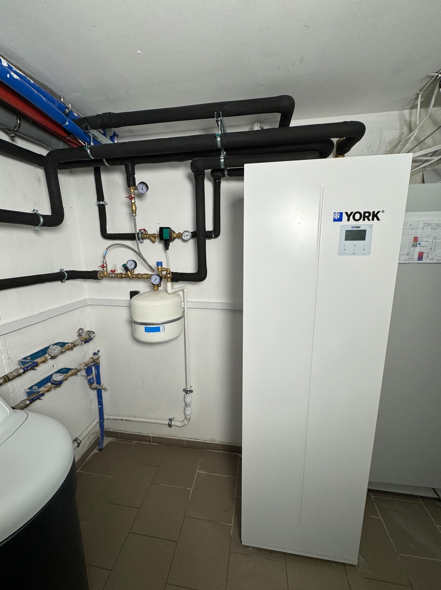 Biała pompa ciepła marki York w kotłowni, widoczne rury instalacyjne z izolacją, manometry i zbiornik wyrównawczy.