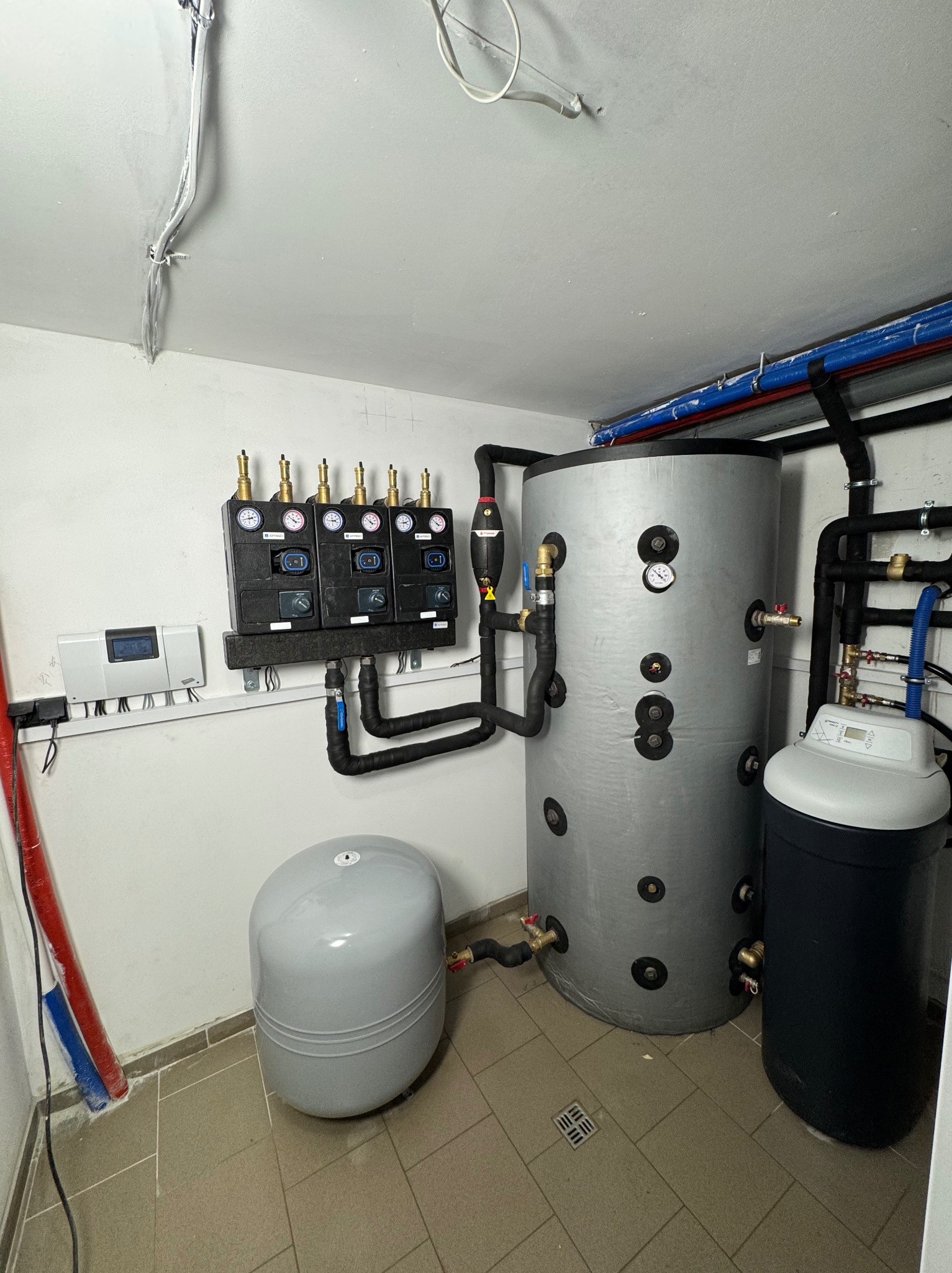 Kotłownia z szarym zasobnikiem ciepłej wody, czarnym filtrem wody, szarym naczyniem wzbiorczym, rozdzielaczem podłogowym i sterownikiem na ścianie, widoczne rury instalacyjne.
