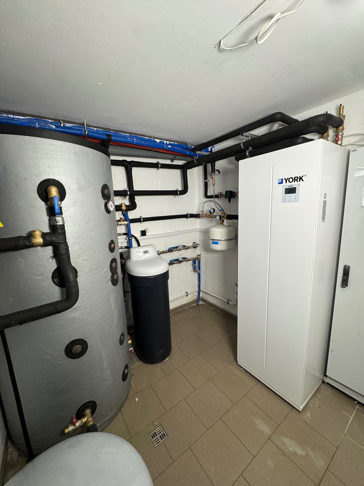 Zestaw instalacji: zbiornik akumulacyjny, zmiękczacz wody i pompa ciepła York w kotłowni z widocznymi rurami instalacyjnymi.