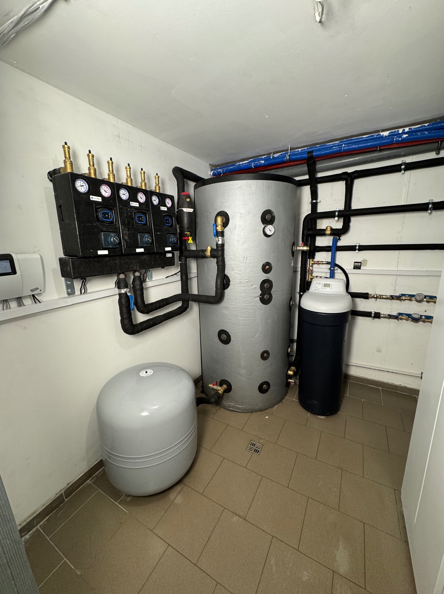 Kotłownia z szarym zbiornikiem akumulacyjnym, czarnym systemem kontroli z manometrami, szarym zbiornikiem wyrównawczym i filtrem wody na brązowych płytkach podłogowych.