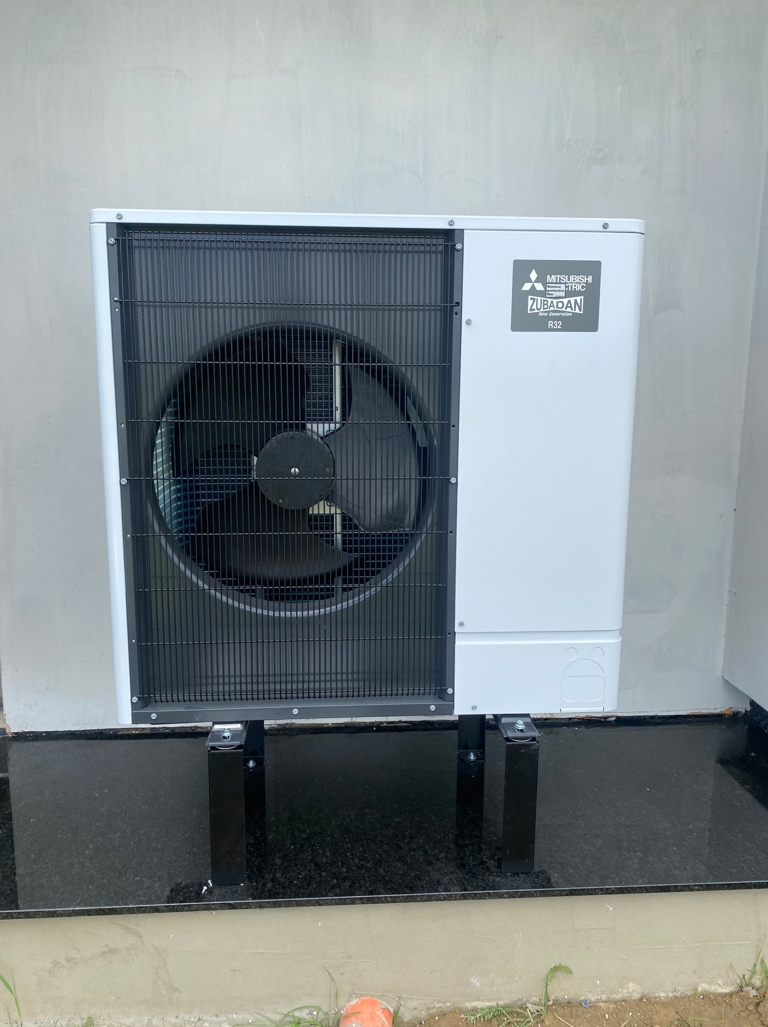 Biała zewnętrzna jednostka pompy ciepła Mitsubishi Zubadan zamontowana na czarnych, metalowych wspornikach na ciemnej, granitowej podstawie przy szarej ścianie.