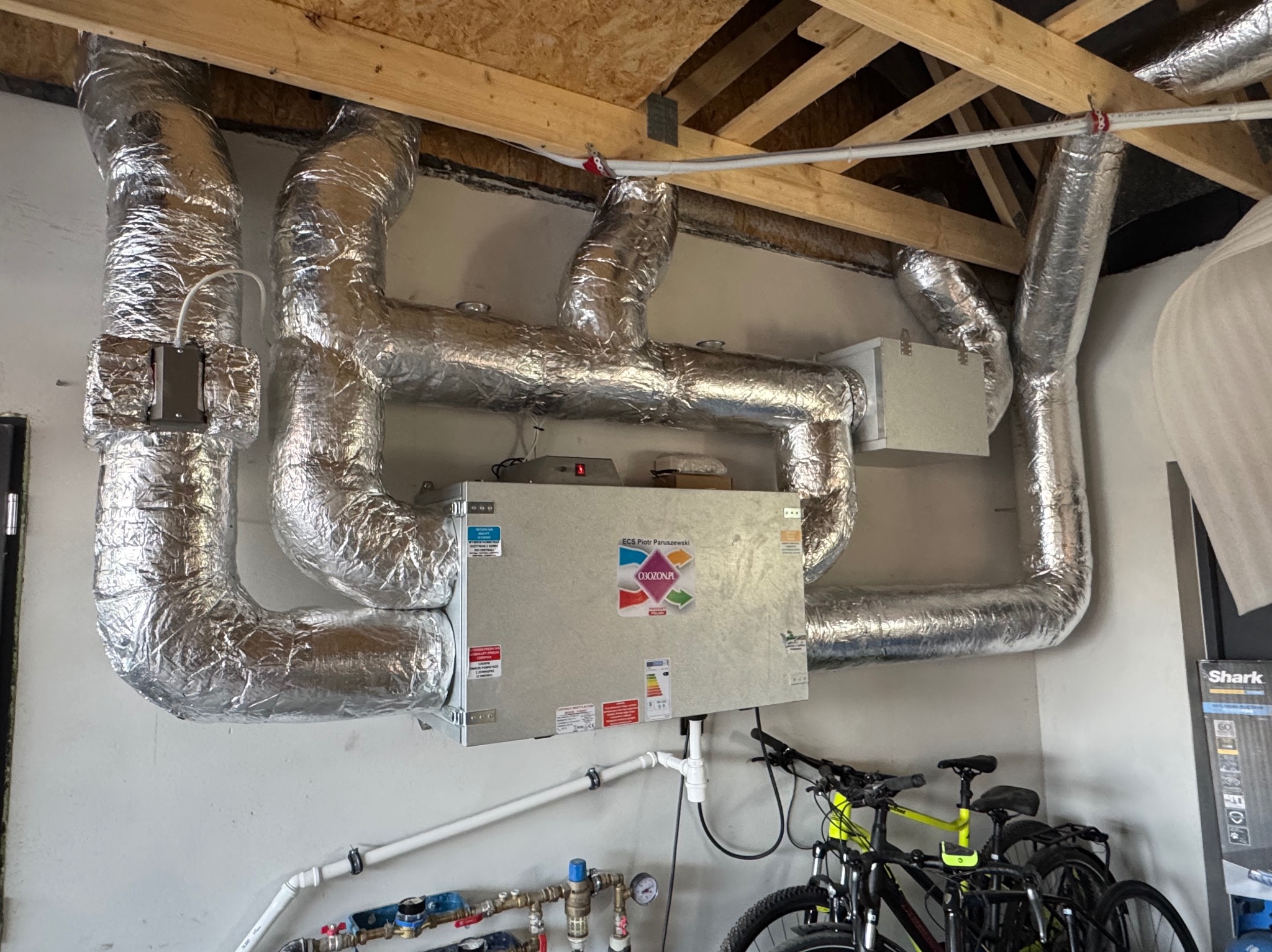 Centrala wentylacyjna z rekuperacją, z widocznymi przewodami izolowanymi folią aluminiową, zamontowana w pomieszczeniu z surowym drewnianym sufitem, z rowerami w tle.