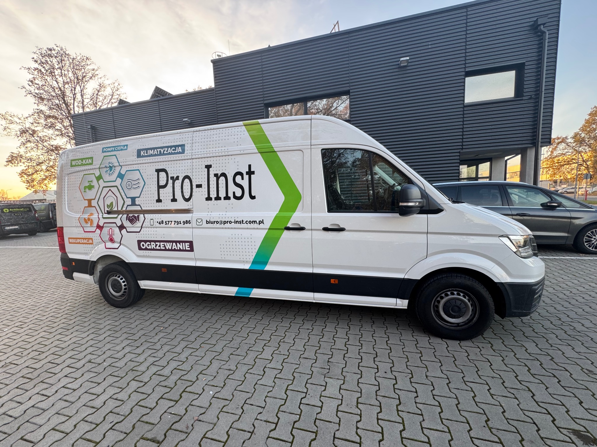Biały samochód dostawczy z logo Pro-Inst i informacjami o usługach: wod-kan, pompy ciepła, klimatyzacja, rekuperacja i ogrzewanie, zaparkowany na brukowanym parkingu przed nowoczesnym budynkiem.