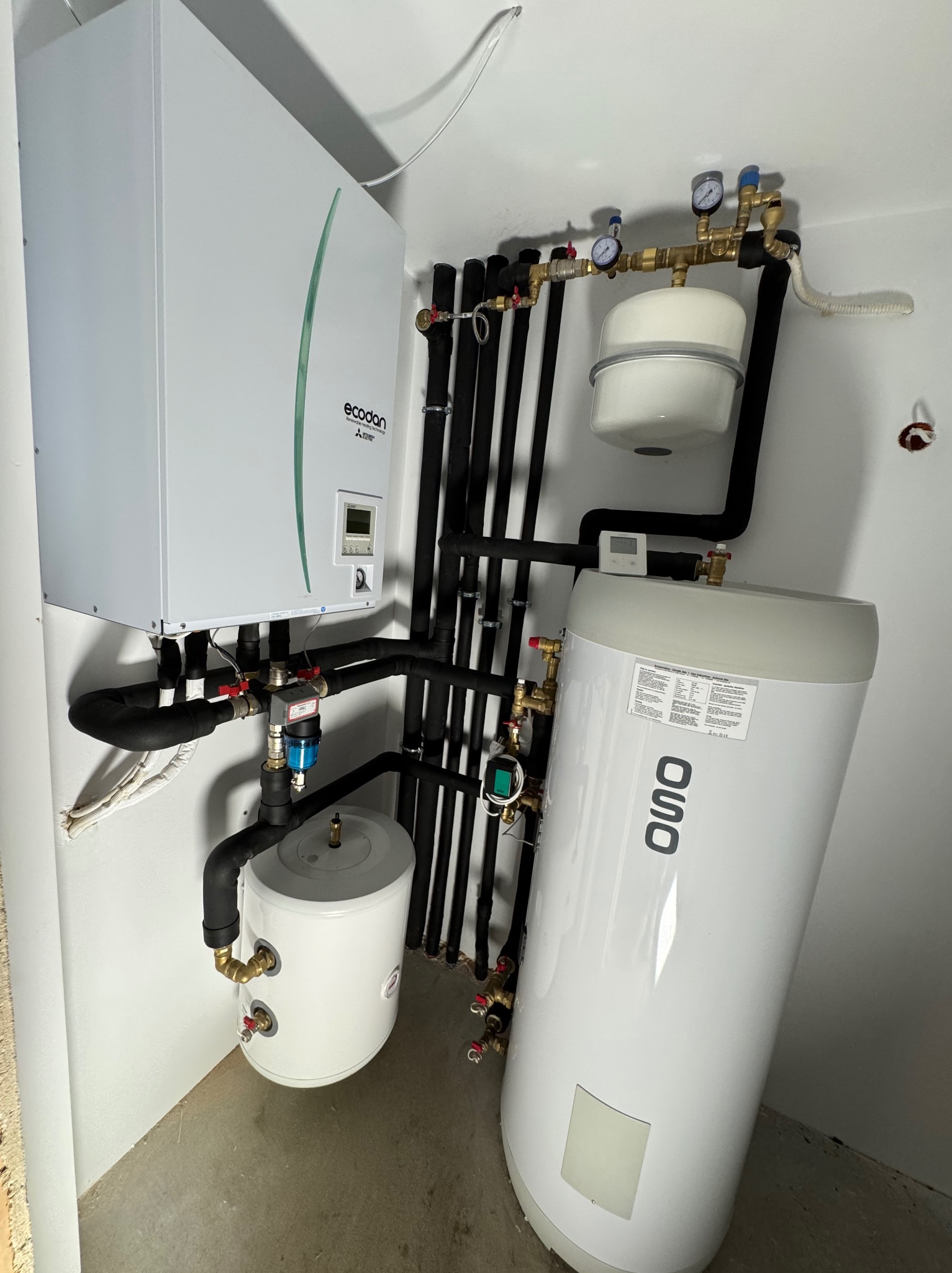 Zestaw urządzeń w kotłowni: biały moduł Ecodan z zielonym akcentem, pionowy zbiornik OSO z czarnym napisem, mniejszy zbiornik, czarne rury izolacyjne, manometry i zawory, betonowa podłoga.