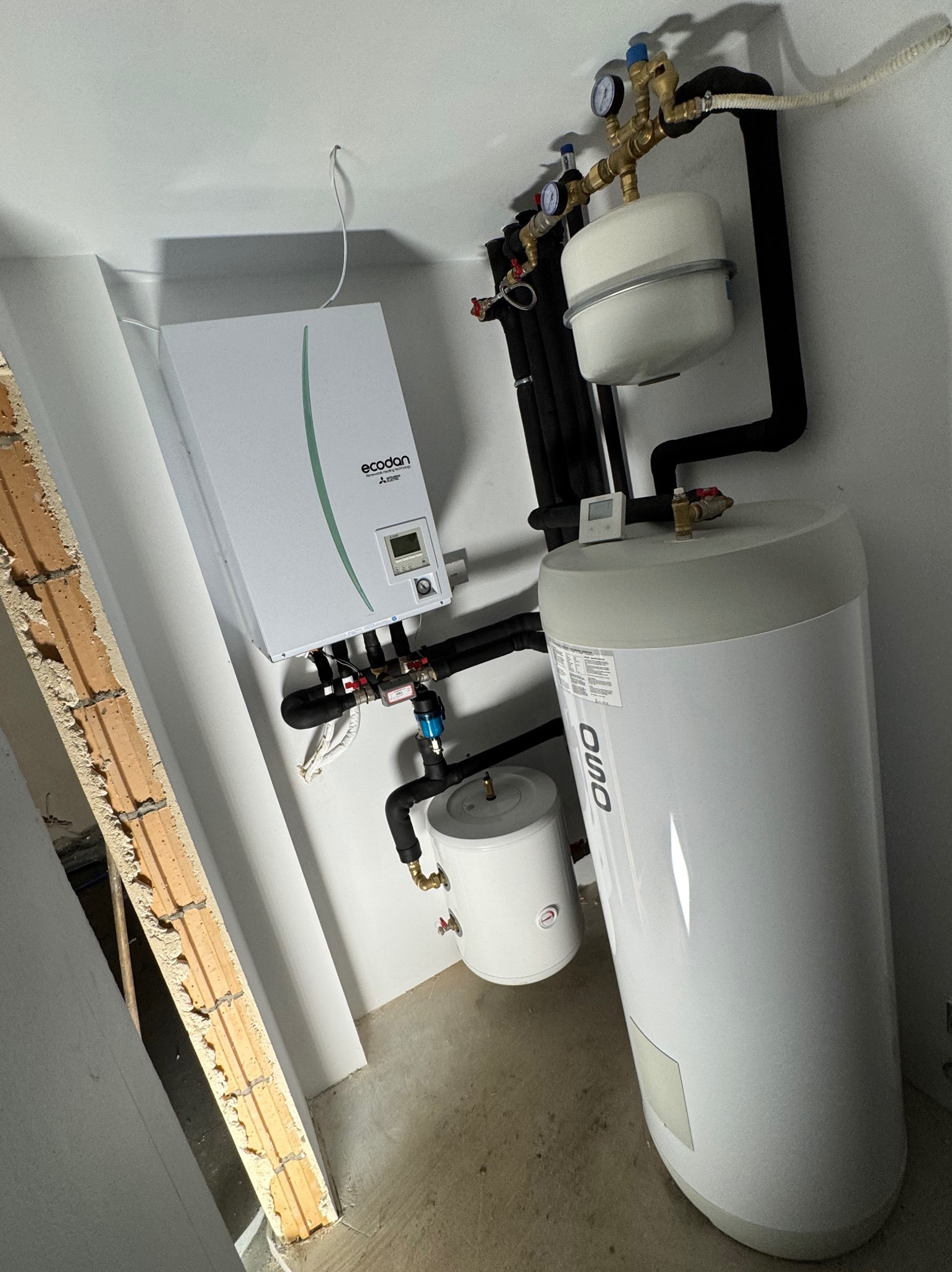Biała pompa ciepła Ecodan Mitsubishi Electric z podłączonymi rurami i zbiornikiem OSO w surowym pomieszczeniu z widoczną ścianą z cegieł.