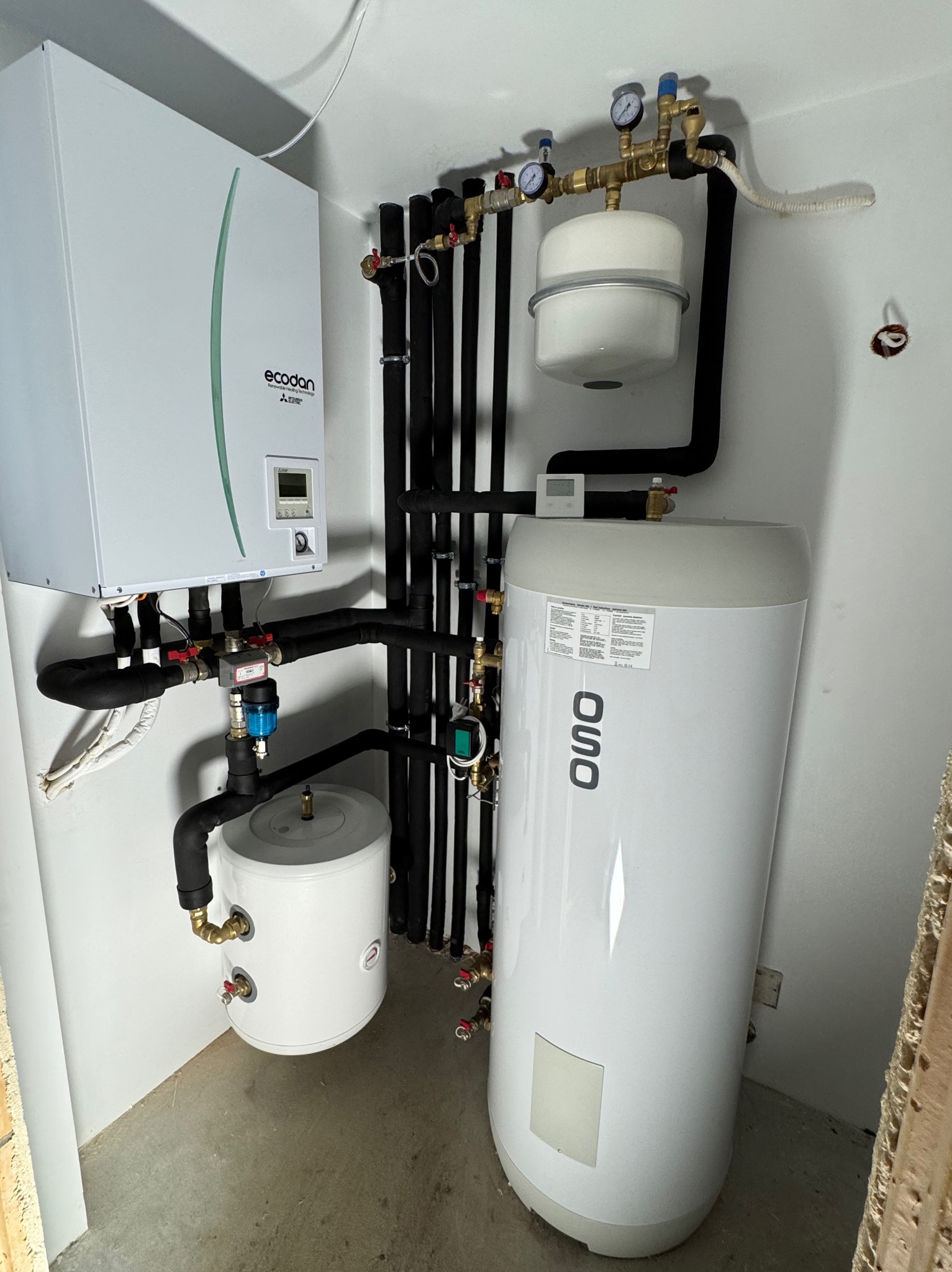 Biała ściana z zamontowaną instalacją centralnego ogrzewania: pompa ciepła Mitsubishi Ecodan, zbiornik buforowy OSO, rury w izolacji termicznej i zawory, widoczne manometry.