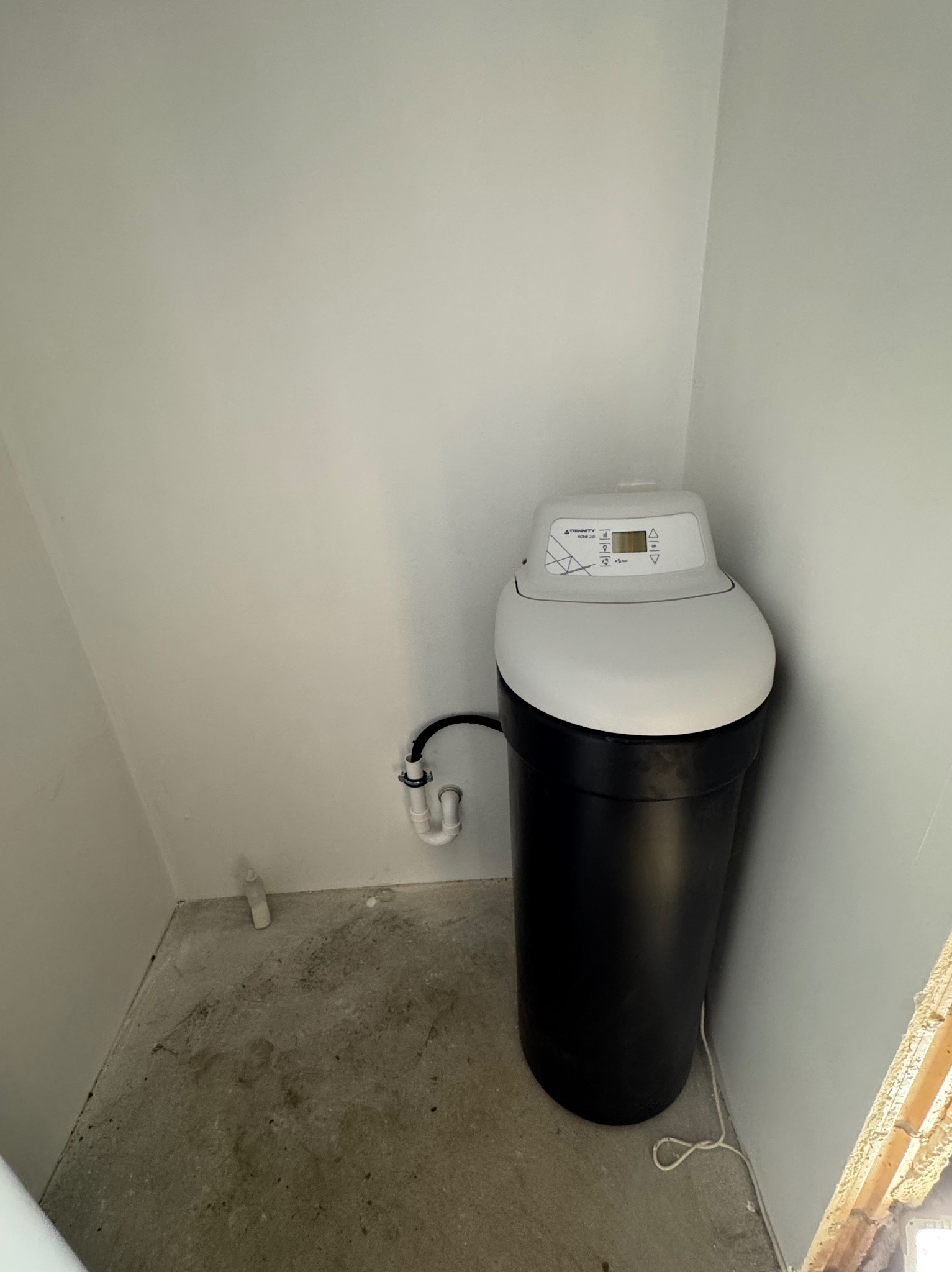 Czarno-biały zmiękczacz wody marki Trinity Home 2.0 zainstalowany w rogu pomieszczenia z surowym betonem na podłodze i widocznymi rurami przyłączeniowymi do ściany.