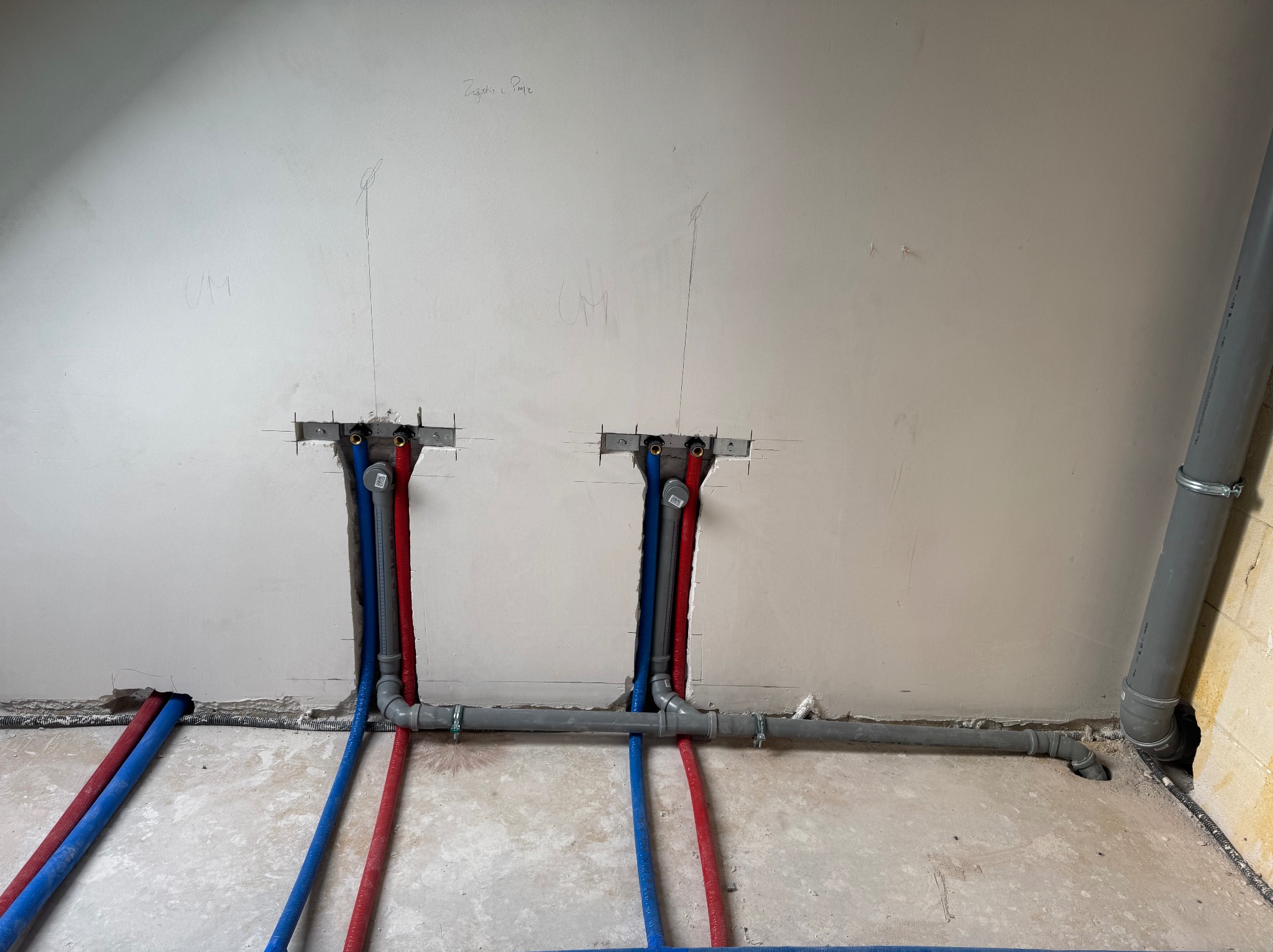 Wewnętrzna instalacja wodna z rurami PEX w kolorze niebieskim i czerwonym, wychodzącymi ze ściany, widoczne mocowania i szare rury kanalizacyjne, surowy stan wykończenia wnętrza.