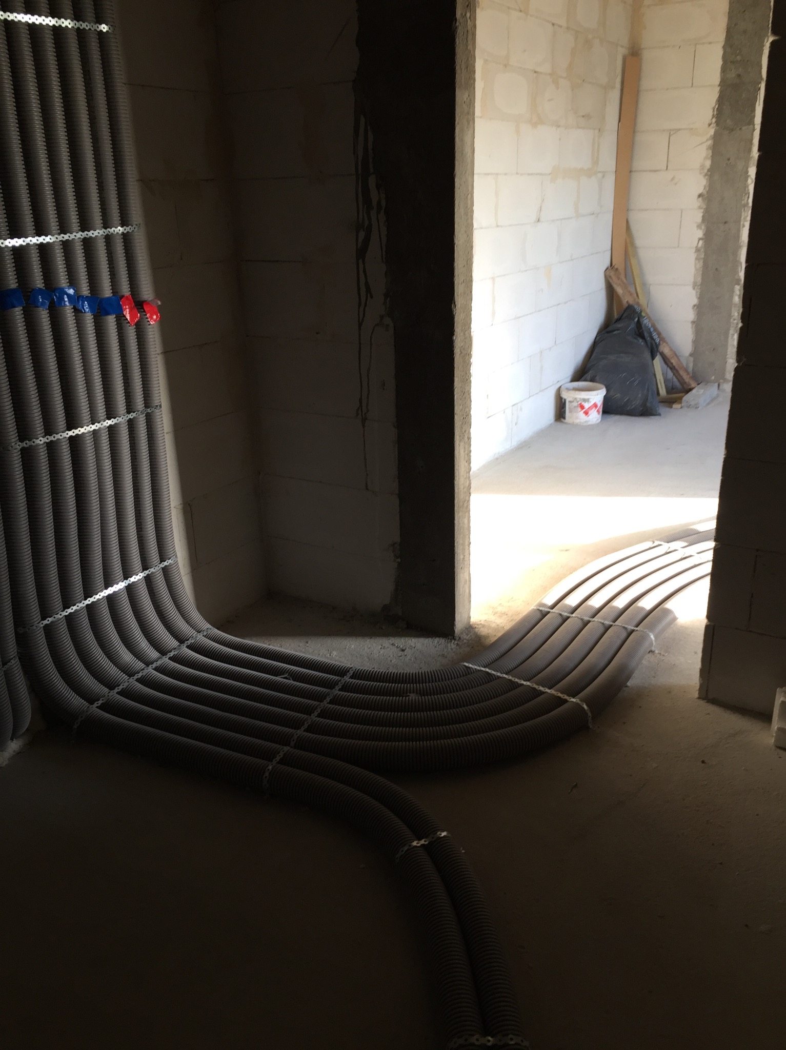 Szare rury instalacyjne, zabezpieczone metalowymi taśmami, prowadzone w surowym wnętrzu budynku z bloczków, widoczne oznaczenia kolorami niebieskim i czerwonym.