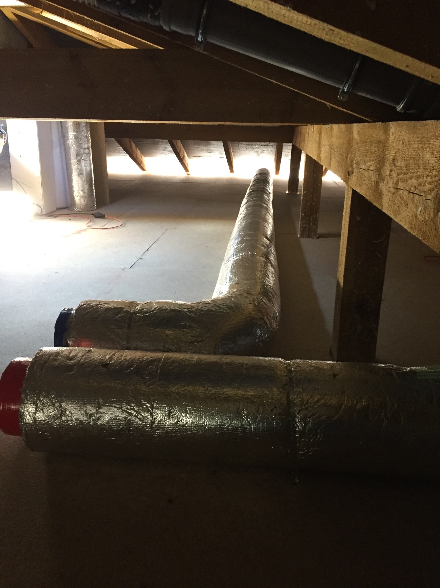 Dwie izolowane rury wentylacyjne ułożone na podłodze pod drewnianą konstrukcją dachu, widoczne mocowania rur do belek, w tle jasne światło.