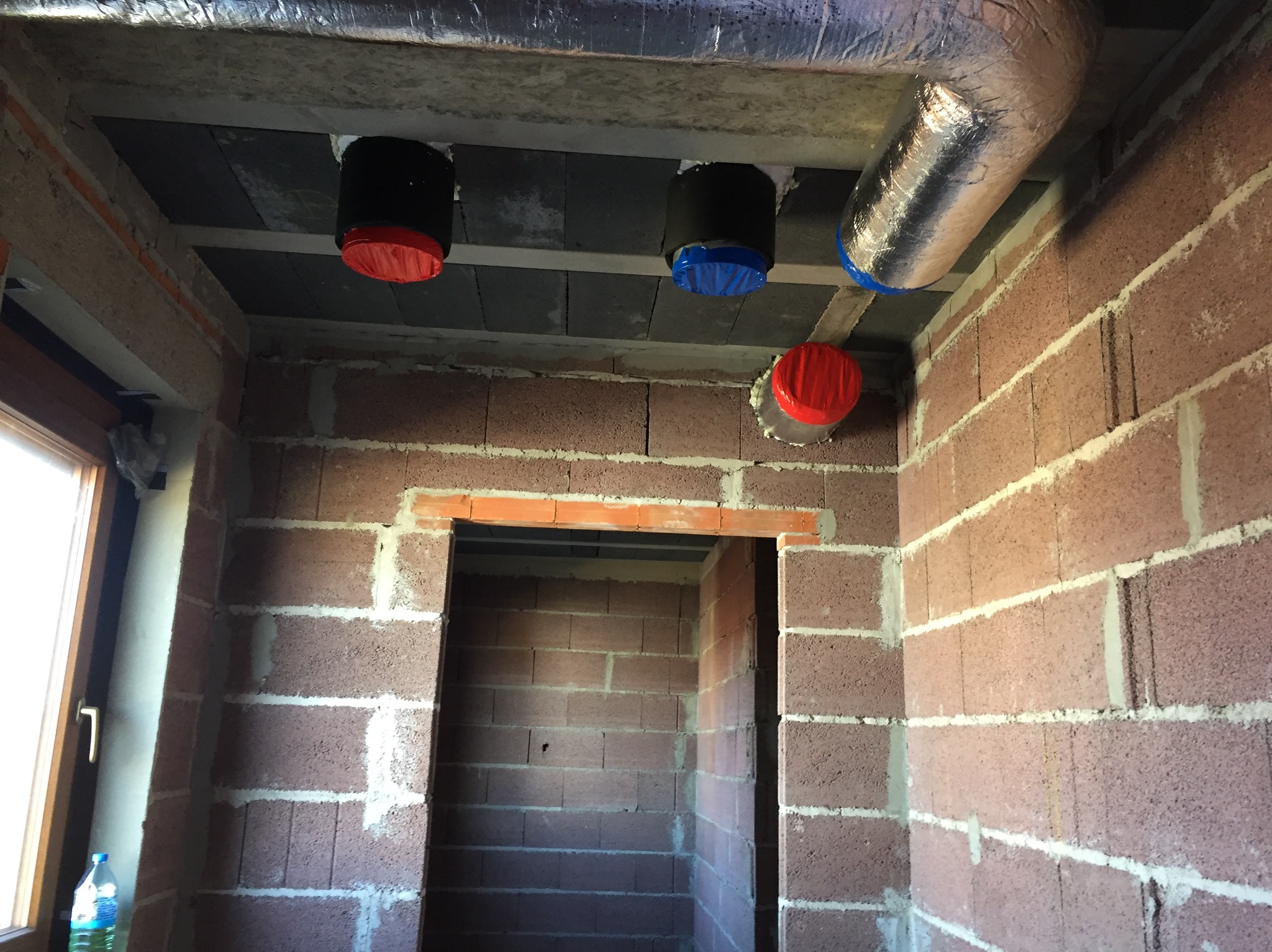 Wnętrze w trakcie montażu rekuperacji: widoczne kanały wentylacyjne z folią aluminiową, czarne rury z czerwonymi i niebieskimi zaślepkami na suficie z betonowych pustaków, ściany z czerwonej cegły...