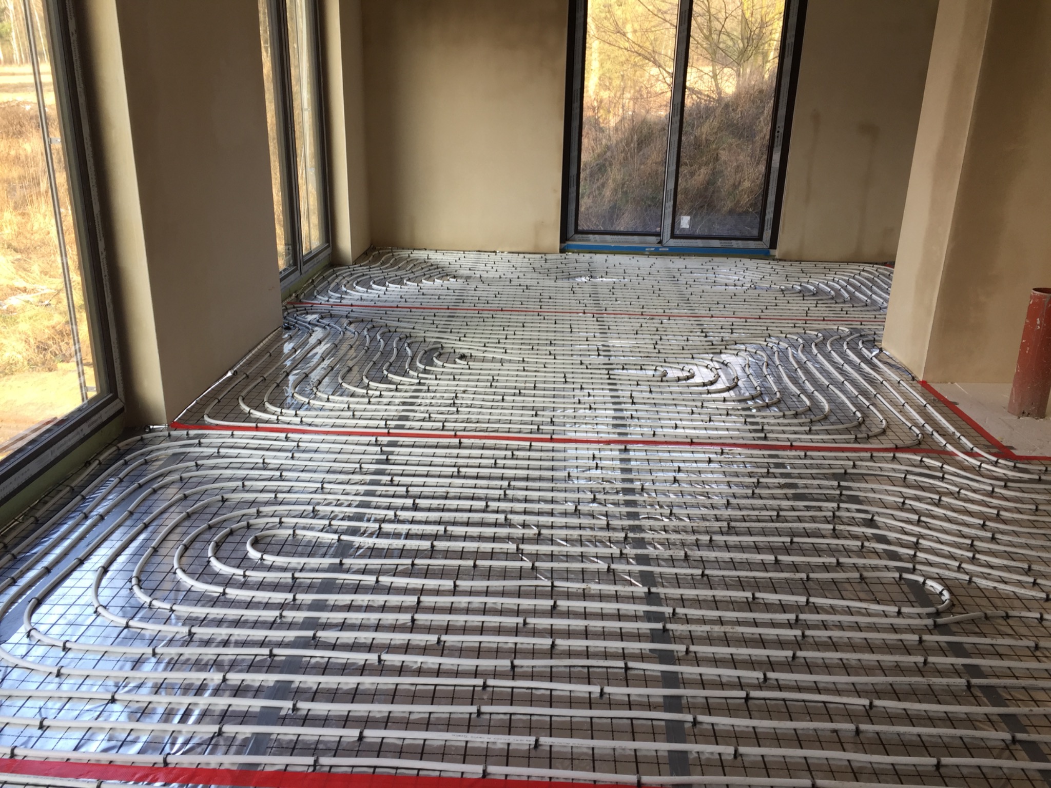 Białe rury grzewcze ułożone w pętle na metalowej siatce pod folią izolacyjną, przygotowane do zalania wylewką w jasnym pomieszczeniu z dużymi oknami.