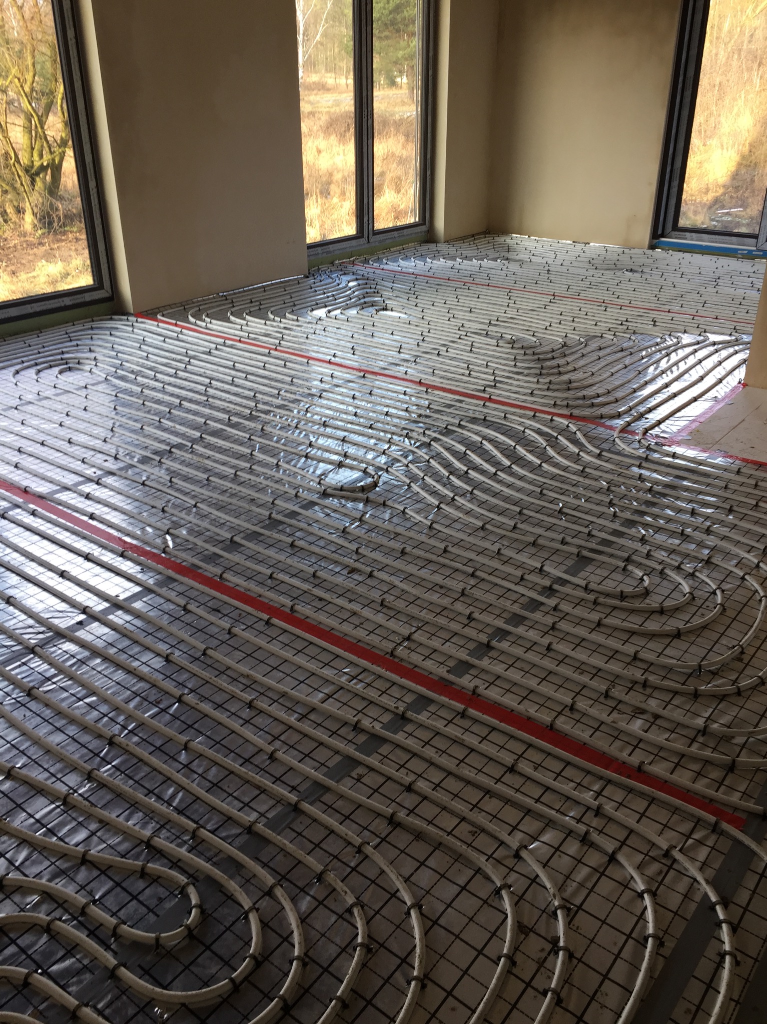 Białe rurki ogrzewania podłogowego ułożone w pętle na czarnej siatce, zabezpieczone folią, z widocznymi czerwonymi taśmami mocującymi w jasnym pomieszczeniu z dużymi oknami.
