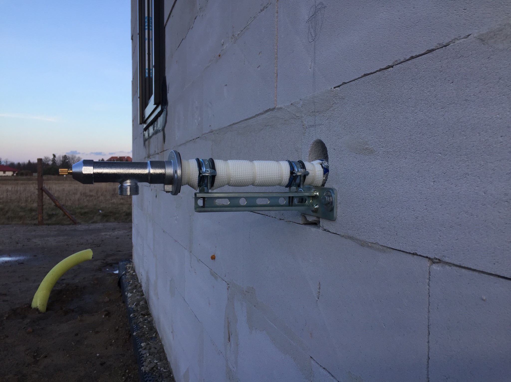 Zewnętrzny zawór z izolacją termiczną zamontowany na ścianie budynku z betonu komórkowego, widoczny fragment okna i wystająca żółta rura w ziemi.