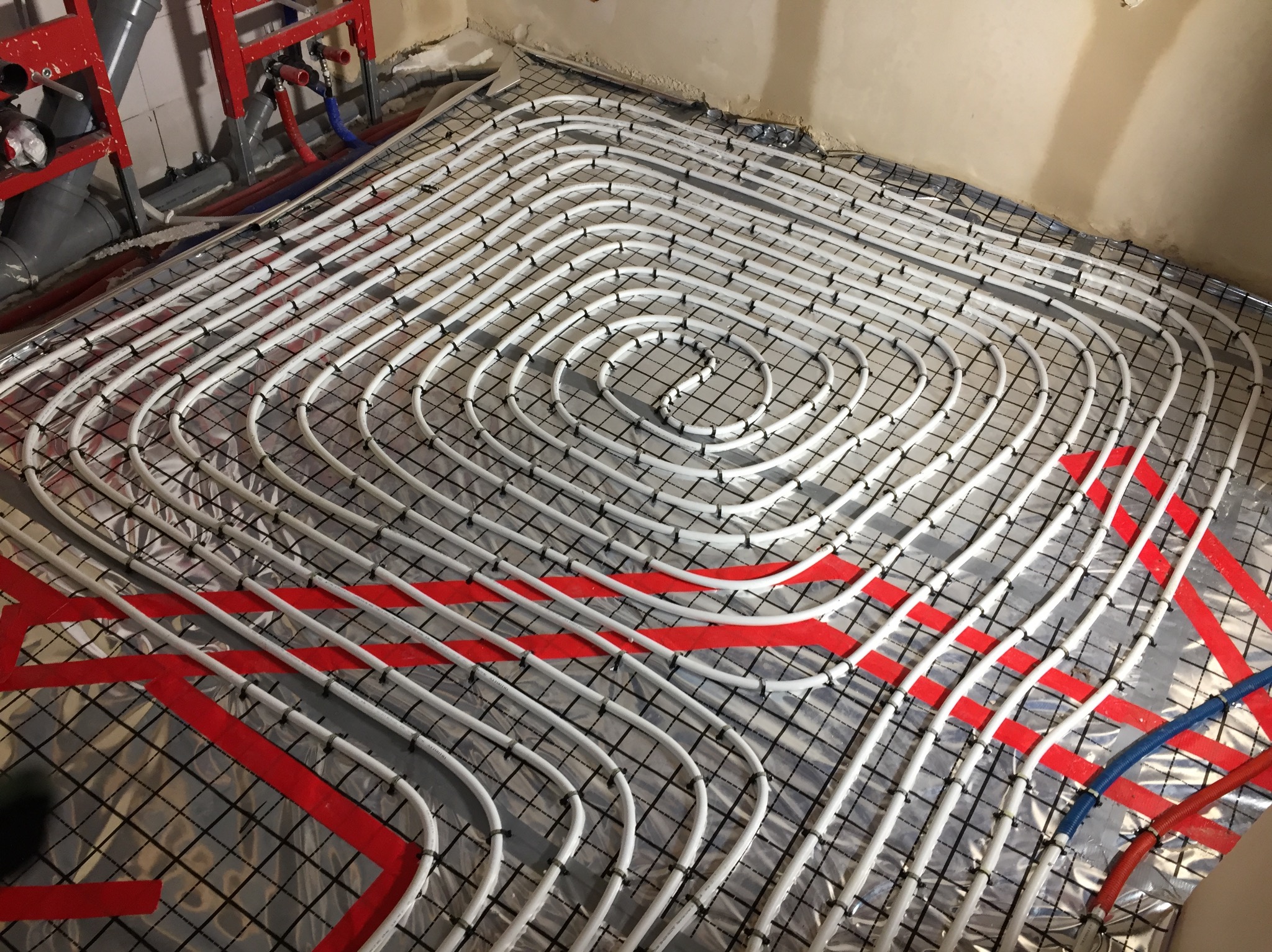 Białe rury systemu ogrzewania podłogowego ułożone spiralnie na czarnej siatce, przymocowane plastikowymi klipsami, z czerwonymi taśmami wyznaczającymi strefy, widoczne fragmenty instalacji...