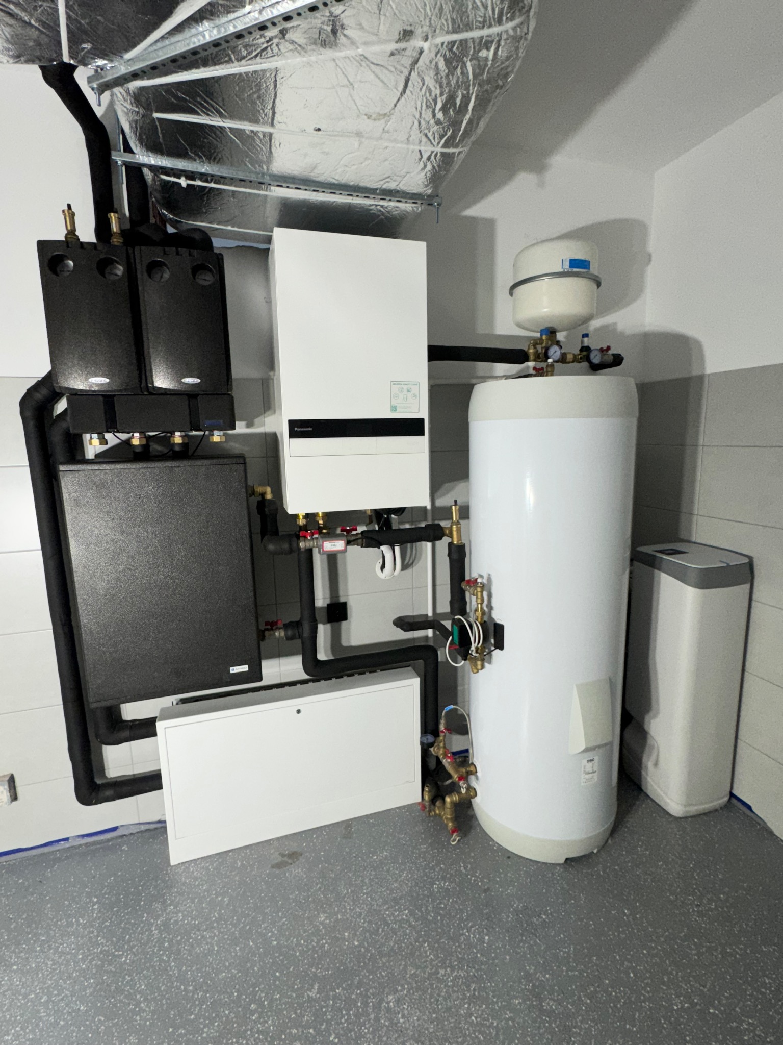 Zestaw urządzeń grzewczych w kotłowni: dwa czarne moduły, biały piec kondensacyjny, biały zasobnik ciepłej wody, szare rury, izolacja na suficie.