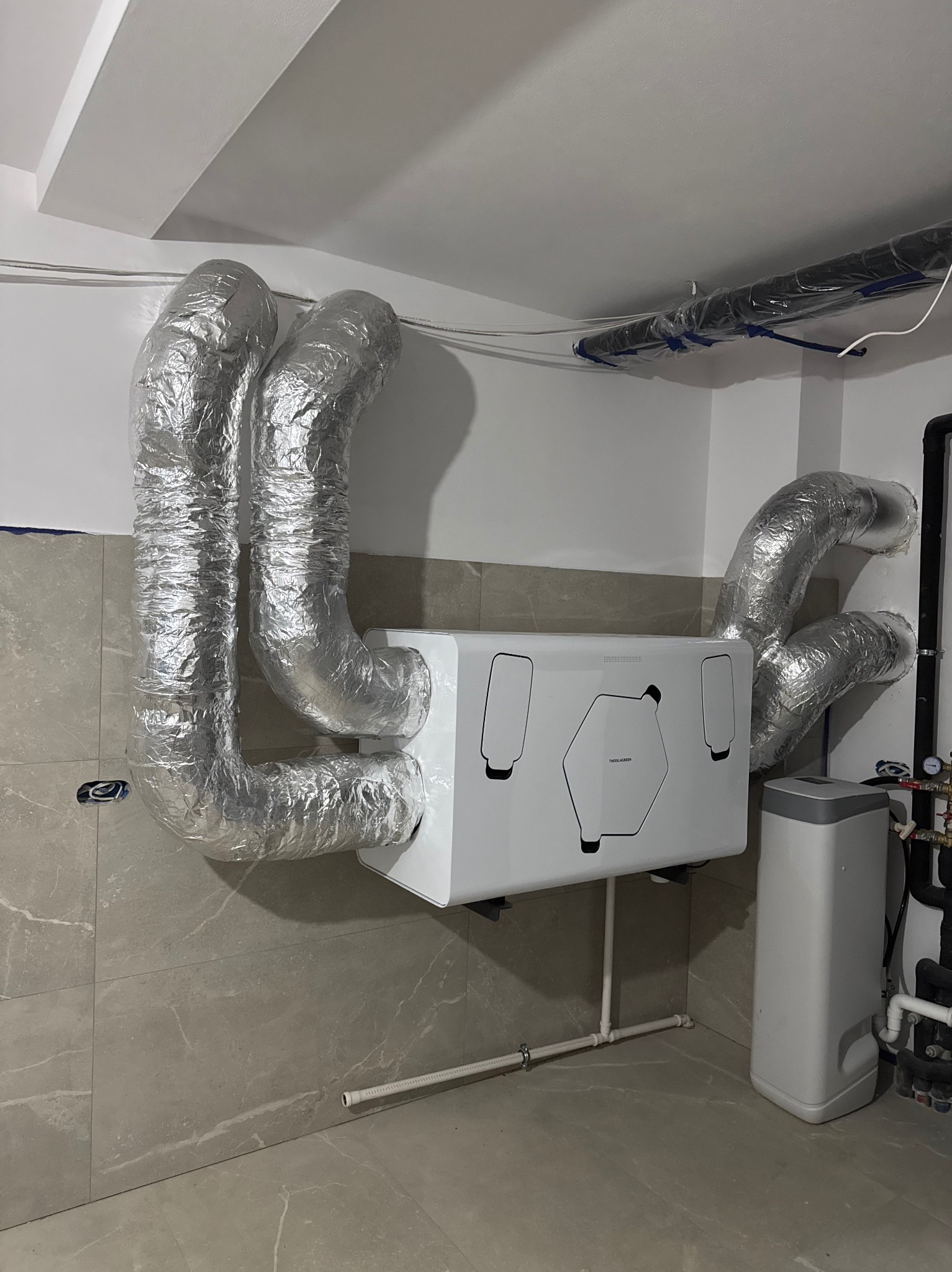 Centrala wentylacyjna z rekuperacją, z widocznymi przewodami izolowanymi folią aluminiową, zamontowana na ścianie z płytek ceramicznych w pomieszczeniu technicznym.