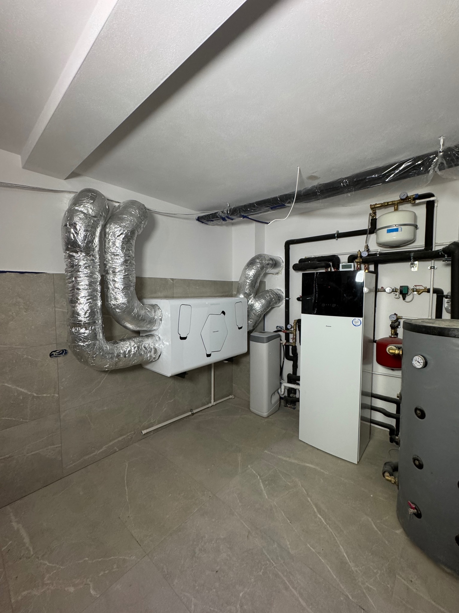 Zainstalowana jednostka rekuperacyjna z aluminiowymi kanałami wentylacyjnymi w pomieszczeniu technicznym z szarą płytką podłogową i kotłem grzewczym po prawej stronie.