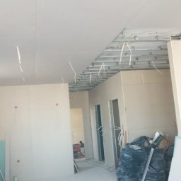 Artur Szypuła - Wnętrze w trakcie montażu płyt kartonowo-gipsowych: widoczne stelaże sufitowe, częściowo zamontowane płyty, kable elektryczne zwisające z sufitu, worki z odpadami budowlanymi i płyty gipsowe oparte...